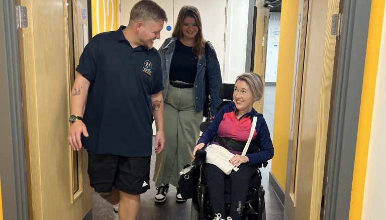 Sarah with a companion meet physiotherapist Dan entering the MoveWell Centre. Sarah uses a wheelchair, Dan is walking next to her and the companion is walking behind. All three are smiling and engaged in conversation.
