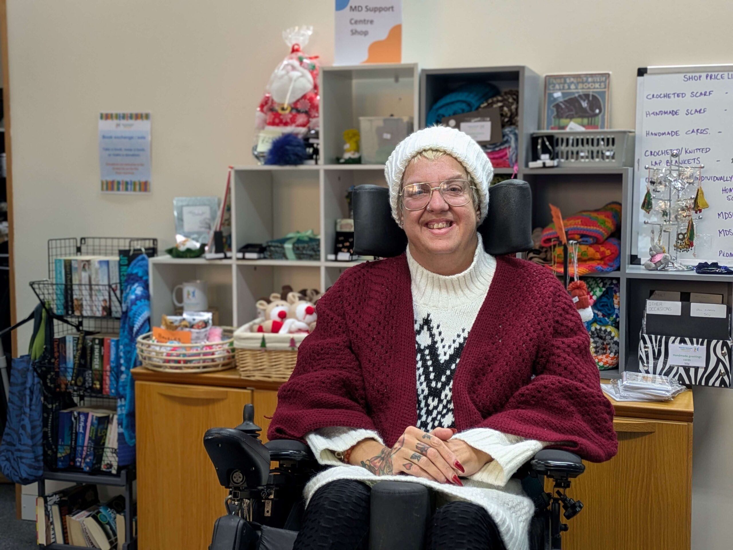 Clair is wearing a red shawl she crocheted herself. She is sitting in her power chair in the MDSC reception and looks happy
