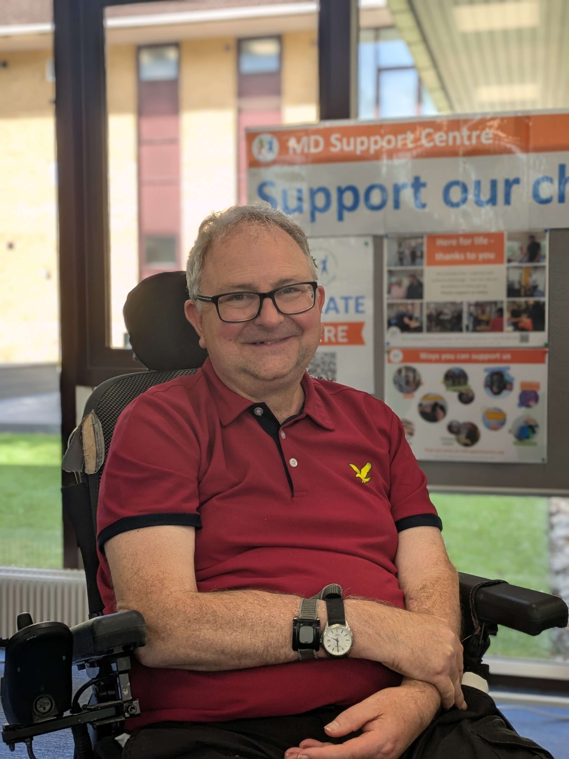 Mark is sitting in his power chair in the MDSC reception. He is smiling at the camera. He's wearing a red t shirt and has his arms folded on his lap.