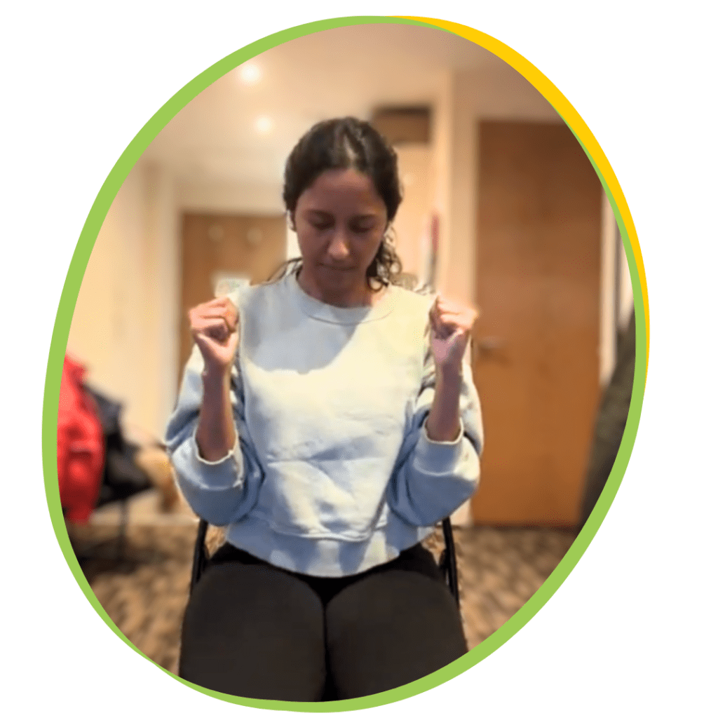 Physiotherapist Rewati demonstrating chair yoga for people with muscular dystrophy. She is wearing a grey or white top, has her arms bent in front of her, and is looking down with her eyes closed.