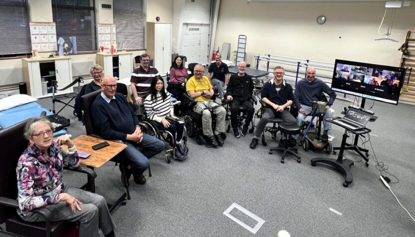 A group of people in chairs and wheelchairs sitting in a semi circle. There is also a TV with people who have joined online.