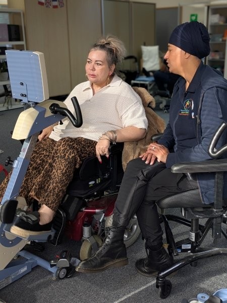A woman uses a wheelchair accessible theratrainer bike while talking to Therapy Assistant Suki