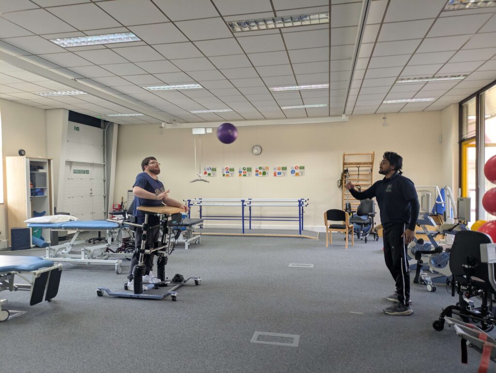 Tommy having a physiotherapy session with Abbas. Tommy is using the Balo standing frame to support him. Abbas has thrown a ball and Tommy is getting ready to hit it.