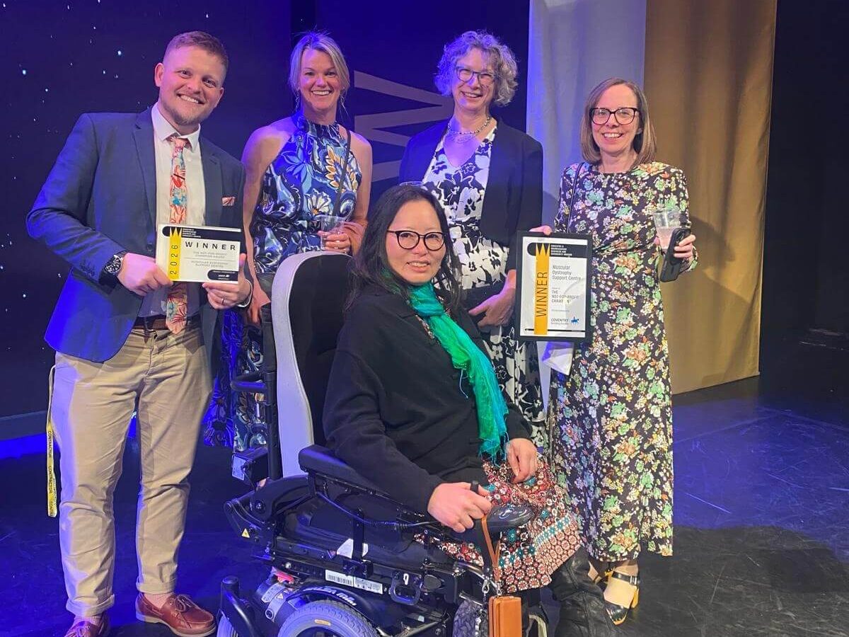 Small group of award winners posing on stage at the Coventry & Warwickshire Business and Community Awards, holding certificates next to an event banner, with one attendee seated in a powered wheelchair.