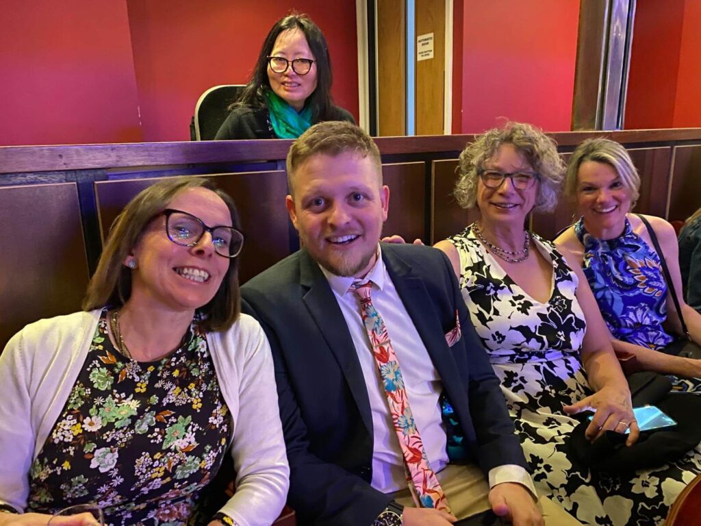 Five attendees seated in a theatre-style venue, dressed formally, looking towards the camera, with red walls and wooden seating visible in the background.