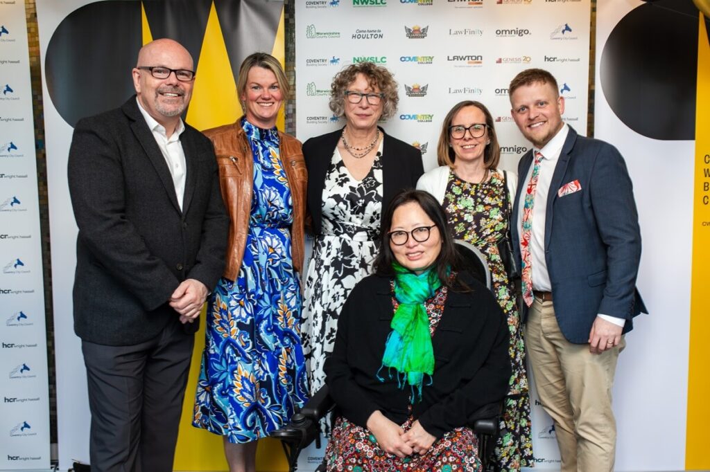 Group photo of award winners standing in front of event branding and black and gold balloons, with certificates on display and one attendee seated in a powered wheelchair at the front.