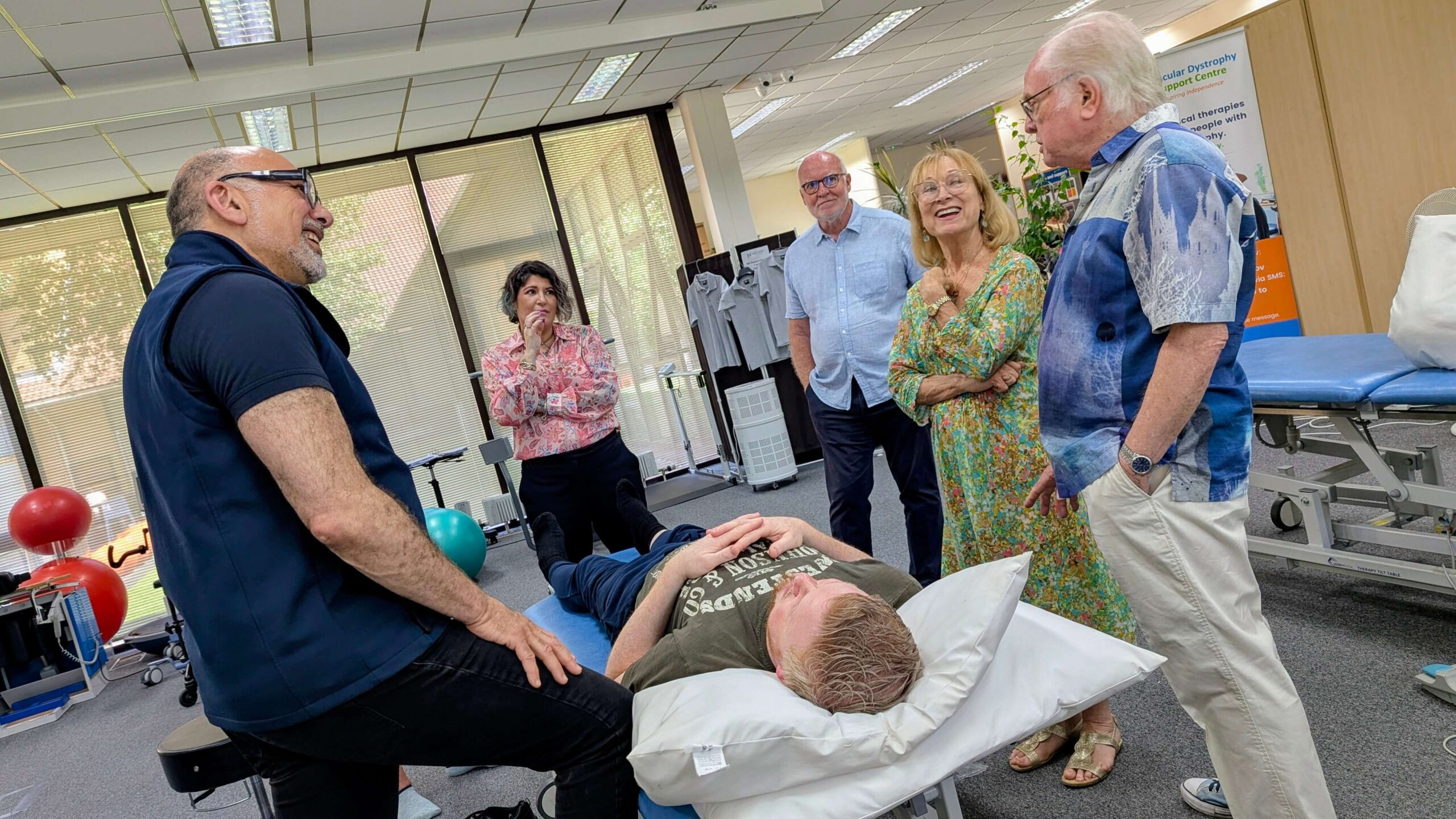 Closer view of a physiotherapy demonstration. A clinician stands beside a person lying on a treatment table, explaining a technique to several adults gathered around. Exercise equipment, windows with blinds, and clinic furnishings are visible behind them.
